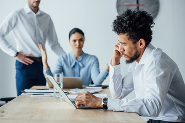 Selective focus of african american man using laptop near angry mexican colleague in office
