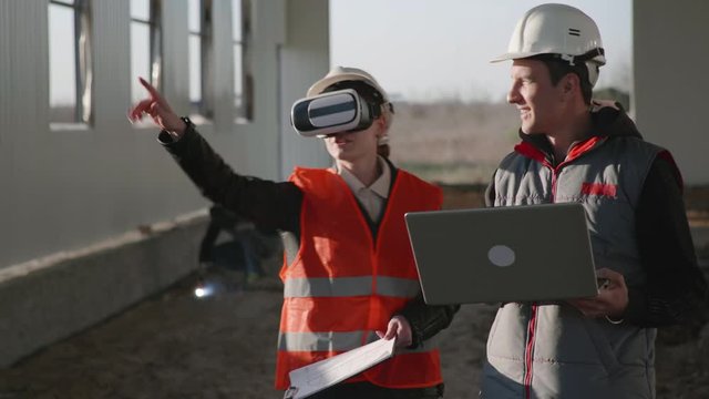 modern technologies, female and male engineer in viar glasses and laptop in hand in protective helmets discuss new building with help of additional reality on construction site