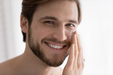 Close up portrait of smiling young caucasian handsome man apply moisturizing face cream or balm for healthy glowing skin, happy millennial male use nourishing beauty product, skincare concept
