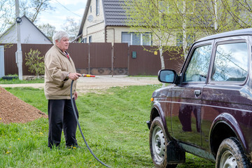 A gray-haired elderly man washes a car outdoors with a garden hose. A powerful jet of water flushes...