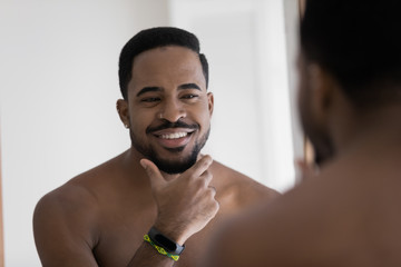 Smiling young african American man look in mirror in bathroom touch shaved bristle or beard, happy biracial millennial male take care do daily beauty facial procedures in bath, skincare cocnept