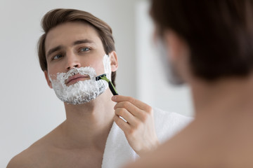 Young caucasian man look in bathroom mirror apply foam gel shaving with razor after shower,...