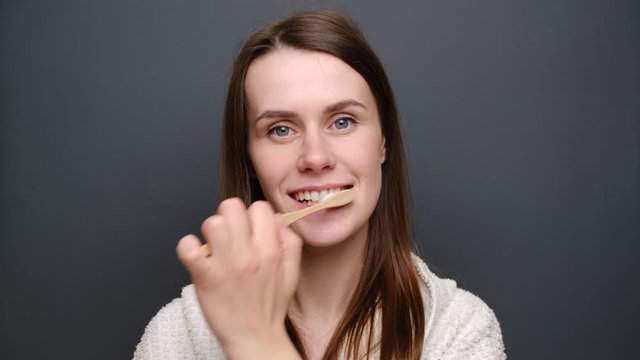 Pretty young woman brushes teeth wooden toothbrush, wears white bathrobe, actively works through all parts of her mouth hygiene on grey studio background. Dental care and reuse concept