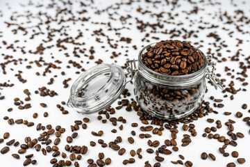 Fototapeta na wymiar Fresh, roasted coffee beans in a open, transparent jar standing on a white table with lots of coffee scattered around.
