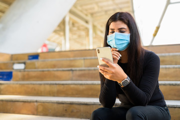 Young Indian woman with mask thinking while using phone by the stairs in city