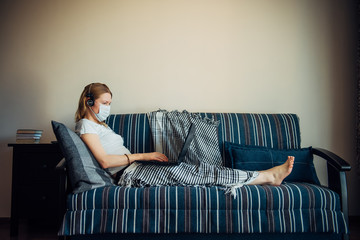 Obraz premium Young caucasian european woman wearing medical mask and plaid using laptop on the sofa during remotely work at home due to coronavirus pandemic.