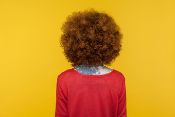 Back view of woman in casual red pullover standing and showing her fluffy curly hair, advertising hairdresser or beauty salon, hair cosmetics, shampoo. indoor studio shot isolated on yellow background