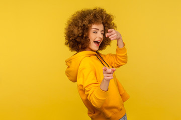 Hey you, handsome! Portrait of joyful curly-haired woman in urban style hoodie winking playfully and pointing to camera, choosing guy and flirting. indoor studio shot isolated on yellow background
