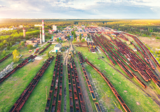 Aerial view of freight trains. Railway station with wagons. Heavy industry. Industrial landscape with train in depot, smoke stack, green trees, buildings, blue sky at sunset. Top view. Transportation