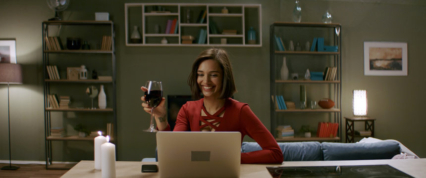 Beautiful woman in a red dress having a glass of wine chatting, online date with partner. Stay home, quarantine life. Shot with anamorphic lens
