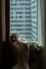 Woman with naked back sitting by a window in flat. Stay isolation at home. Quarantine concept. COVID-19 Pandemic Coronavirus. Residential community. Apartment Building. Social Distancing