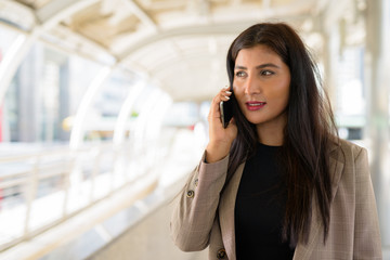 Happy young beautiful Indian businesswoman talking on the phone at footbridge in the city