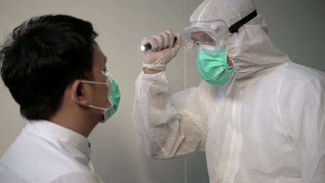 Coronavirus concept. Corona Virus concept. Asian male doctor in a protective suit, protective mask and medical glasses examines a man with a flashlight. 4K Resolution.