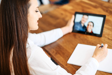 Video consultation, video appointment with family psychologist, doctor. A woman in white shirt is communicating via video with a couple on the tablet screen and doing notes in notebook