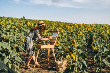 A young woman with curly hair and wearing a hat is painting in nature. A woman stands in a...