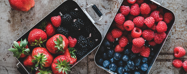 Healthy summer vegan lunch. Flat-lay of fresh seasonal strawberries, raspberries, blueberries and blackberries in lunchboxes over grey background, top view, wide composition. Vegetarian, detox food