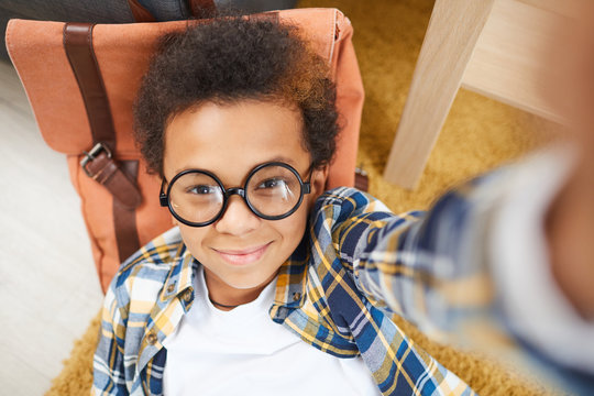 Above view selfie portrait of cute African boy wearing glasses lying on floor next to backpack , copy space