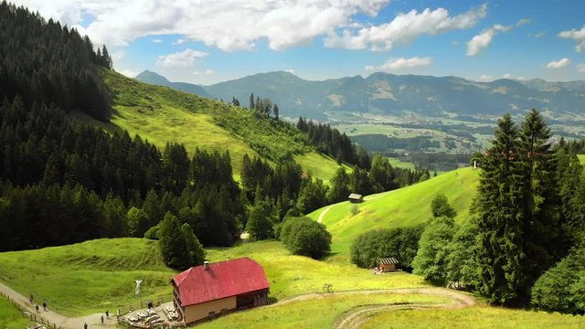 Flying over beautiful mountains with the view to the valley, Alpen in Allgäu, Deutschland,  with hikers walking on green meadows to an inn, enjoying their recreational nature vacation 