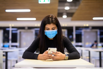 Young Indian woman with mask using phone and sitting with distance at food court