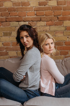 Side View Of Mother And Daughter Looking At Camera While Sitting Back To Back On Couch