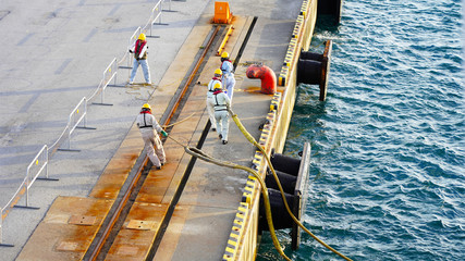 Fototapeta na wymiar Parking mooring of the vessel in the port terminal is carried out by workers in helmets and life jackets, a group of people pull a rope to the shore of the seaport. Passenger terminal in Japan