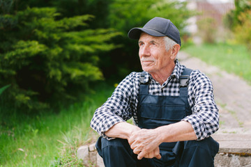 Senior gardener sitting on the steps in garden. Aged worker in overall and baseball cap resting after work. Portrait of old farmer