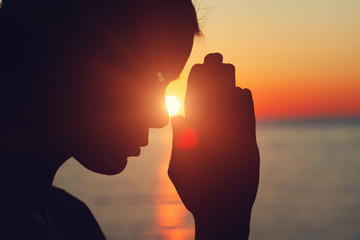 silhouette of young woman raising hands praying at sunset or sunrise light, practicing yoga on the...