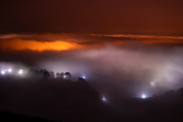 Mountains in fog at beautiful night in autumn in Dalat city, Vietnam. Landscape with Langbiang mountain valley, low clouds, forest, colorful sky with stars, city illumination at dusk.
