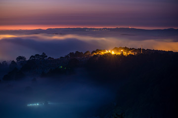 Mountains in fog at beautiful night in autumn in Dalat city, Vietnam. Landscape with Langbiang mountain valley, low clouds, forest, colorful sky with stars, city illumination at dusk.