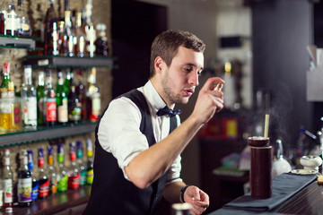 A young guy working as a bartender behind a bar is preparing drinks for customers.