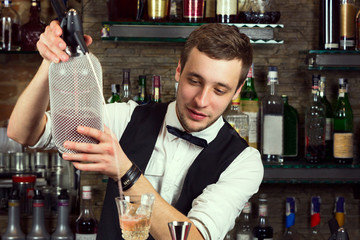 A young guy working as a bartender behind a bar is preparing drinks for customers.