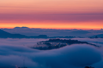 Mountains in fog at beautiful morning in autumn. Landscape with Langbiang mountain valley, low clouds, forest, colorful sky , city illumination at dusk.