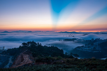 Fototapeta premium Mountains in fog at beautiful morning in autumn. Landscape with Langbiang mountain valley, low clouds, forest, colorful sky , city illumination at dusk.