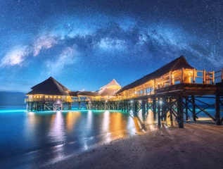 Foto op Canvas Milky Way over wooden bungalow on the water in summer starry night. Landscape with hotel on the sea, illumination, jetty, sandy beach, sky with stars, reflection in water in Zanzibar, Africa. Space © den-belitsky