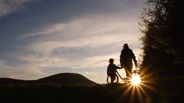Rear view of silhouette of mother with preschool daughter holding hands walking together to bike on a background of mountains during sunset. Сoncept of active family recreation and travel outdoors