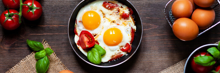 Delicious and healthy breakfast for one person: small pan with 2 fried eggs with vegetables and cheese. Served on a dark wooden table with yellow napkin, fresh tomatoes and basil, brown bread. Banner
