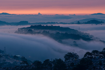 Mountains in fog at beautiful morning in autumn. Landscape with Langbiang mountain valley, low clouds, forest, colorful sky , city illumination at dusk.