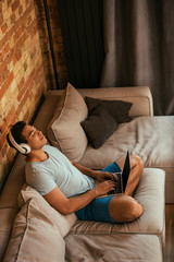 Obraz premium young mixed race man with closed eyes chilling and listening music with headphones and laptop on sofa during self isolation