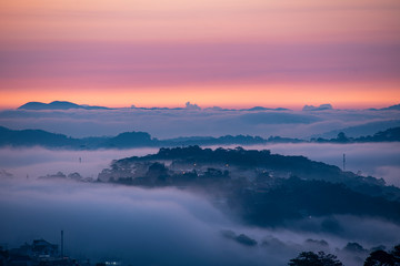 Mountains in fog at beautiful morning in autumn. Landscape with Langbiang mountain valley, low clouds, forest, colorful sky , city illumination at dusk.