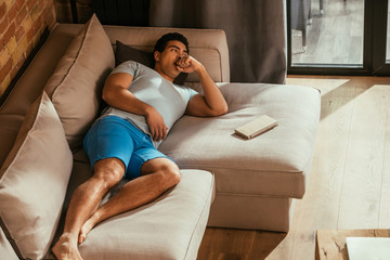 Obraz premium handsome mixed race man lying on sofa with book at home during quarantine
