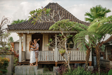 Young lovely couple wearing natural clothes standing on the porch of wooden house, hugs and smiles. Honeymoon on tropical island Bali near wooden bungalow