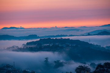 Mountains in fog at beautiful morning in autumn. Landscape with Langbiang mountain valley, low clouds, forest, colorful sky , city illumination at dusk.