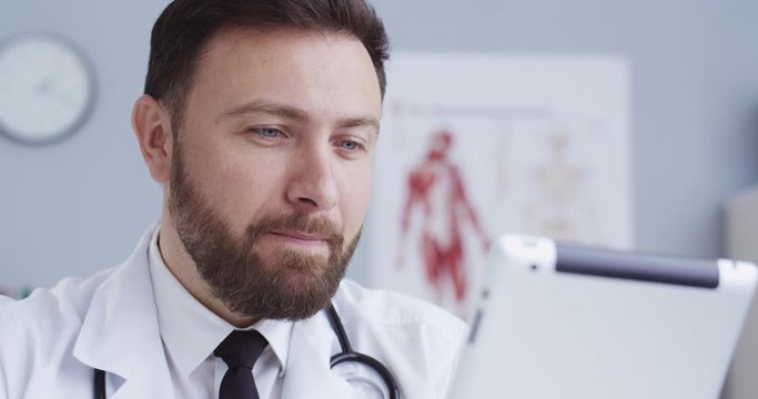 Close-up portrait of family therapist in office in white gown with statoscope speaking using tablet. Doctor consulting online via webcam during quarantine. Сoncept of modern medicine.