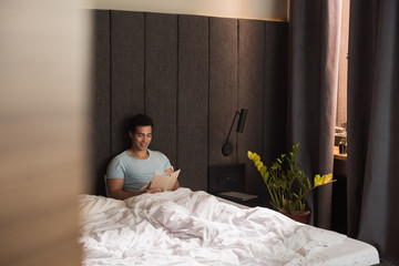 Fototapeta na wymiar happy mixed race man reading book in bed during self isolation, selective focus