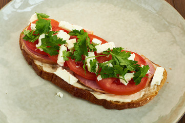 Sandwich of toast and rye bread with greens, tomatoes, feta cheese and ham in grey plate on dark wooden background.