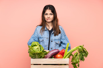 Young farmer woman holding a basket full of fresh vegetables having doubts and with confuse face expression