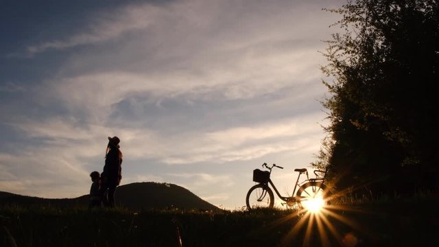 Silhouette of cute little daughter holding hand young mother in hat walking to field on background of mountains during a beautiful sunset. Family on bicycle spends an active weekend in nature