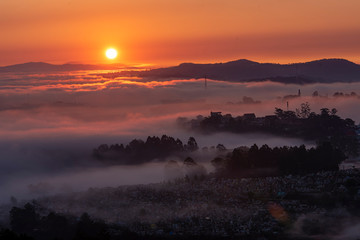 Mountains in fog at beautiful morning in autumn in Dalat city, Vietnam. Landscape with Langbiang mountain valley, low clouds, forest, colorful sky , city illumination at dusk.