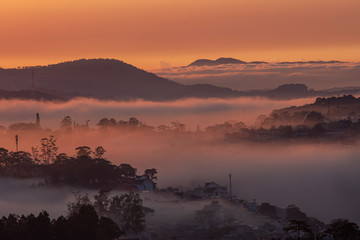 Mountains in fog at beautiful morning in autumn in Dalat city, Vietnam. Landscape with Langbiang mountain valley, low clouds, forest, colorful sky , city illumination at dusk.