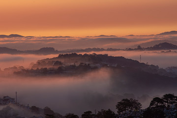Obraz premium Mountains in fog at beautiful morning in autumn in Dalat city, Vietnam. Landscape with Langbiang mountain valley, low clouds, forest, colorful sky , city illumination at dusk.